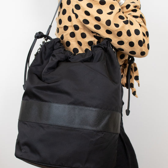 Black nylon drawstring bucket bag displayed on a woman's arm wearing a polka dot top and black pants and standing against a plain background