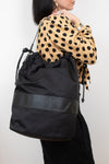 Black nylon drawstring bucket bag displayed on a woman's arm wearing a polka dot top and black pants and standing against a plain background