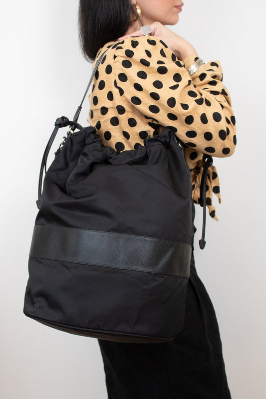 Black nylon drawstring bucket bag displayed on a woman's arm wearing a polka dot top and black pants and standing against a plain background