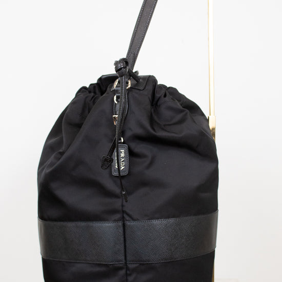Side view of black nylon drawstring bucket bag displayed on a gold stand and against a plain background