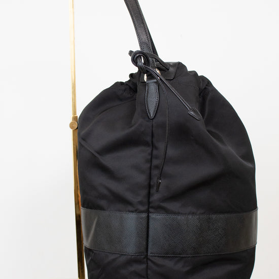 Side view of black nylon drawstring bucket bag displayed on a gold stand and against a plain background