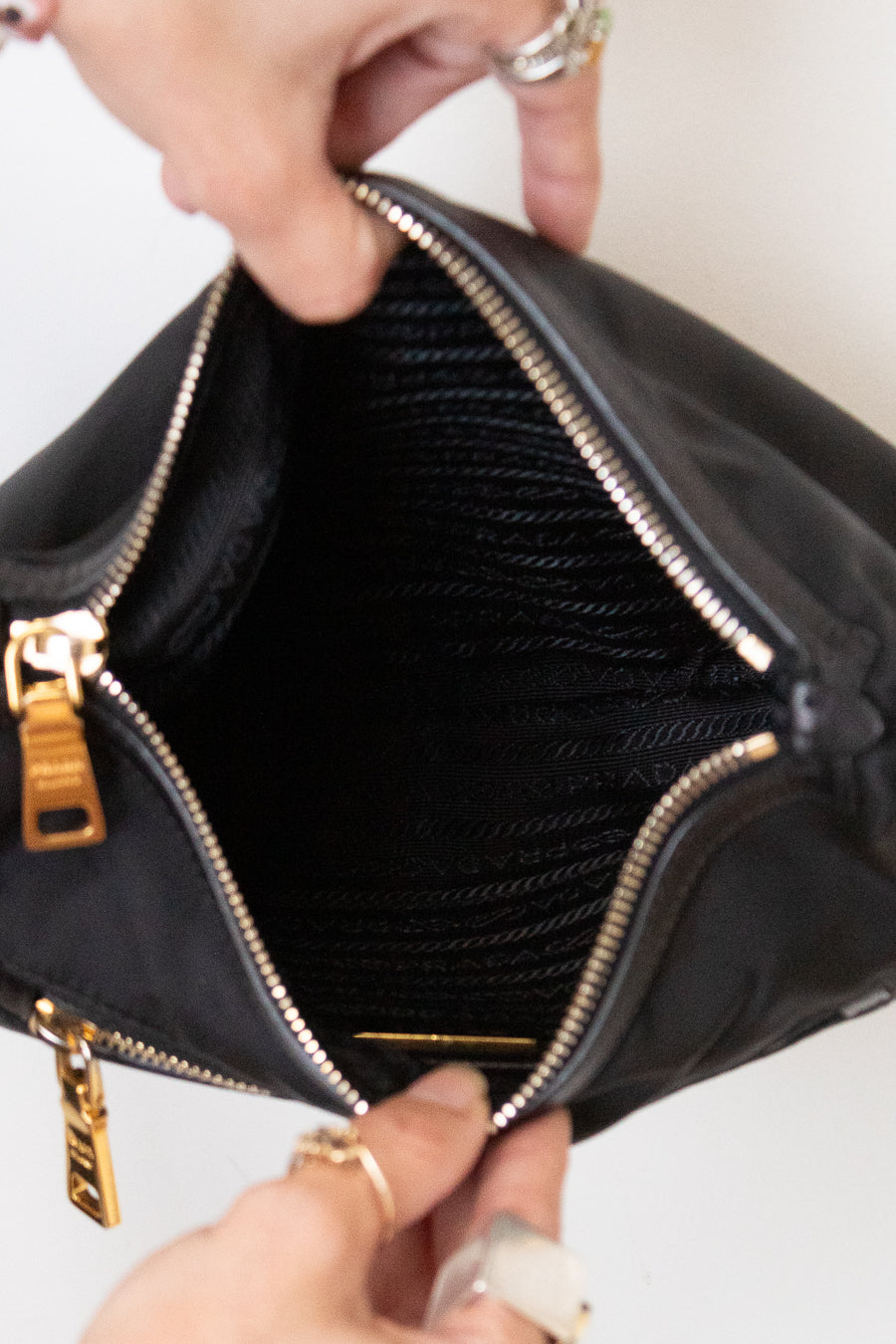 A close-up of a black nylon handbag being opened by two hands wearing silver rings, showing a black mesh interior and gold-tone zippers.