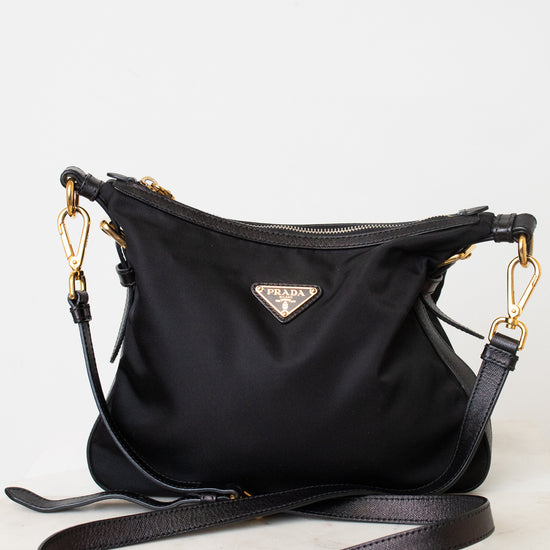 A black Prada nylon shoulder bag with gold-tone hardware and a triangular Prada logo plaque on the front, displayed on a light gray background with its strap draped in front.