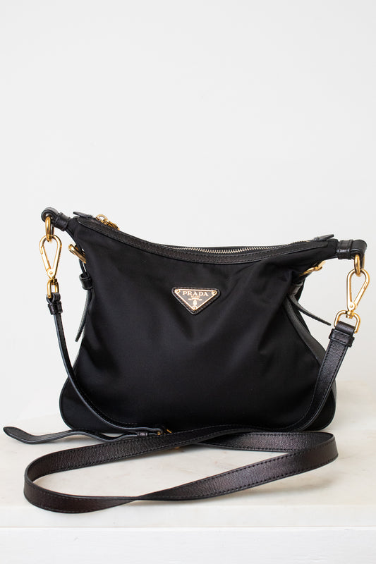 A black Prada nylon shoulder bag with gold-tone hardware and a triangular Prada logo plaque on the front, displayed on a light gray background with its strap draped in front.