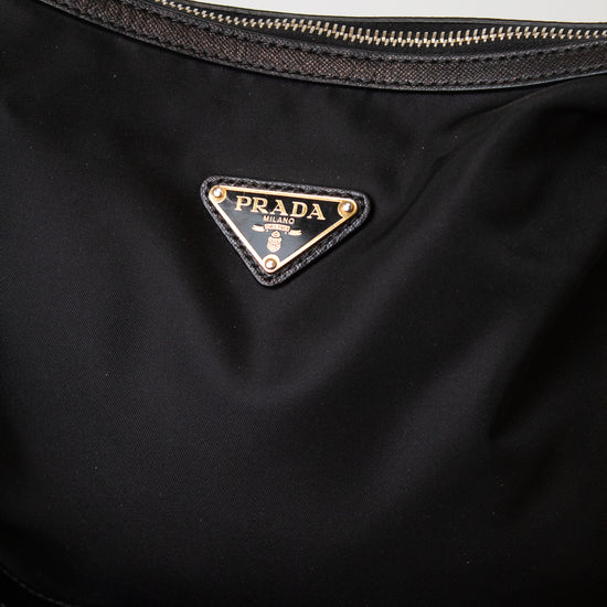 Close-up of a black Prada nylon bag with the iconic triangular Prada logo plaque in gold-tone metal on the front, showing a portion of the black zipper and strap against a light gray background.