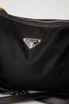 Close-up of a black Prada nylon bag with the iconic triangular Prada logo plaque in gold-tone metal on the front, showing a portion of the black zipper and strap against a light gray background.