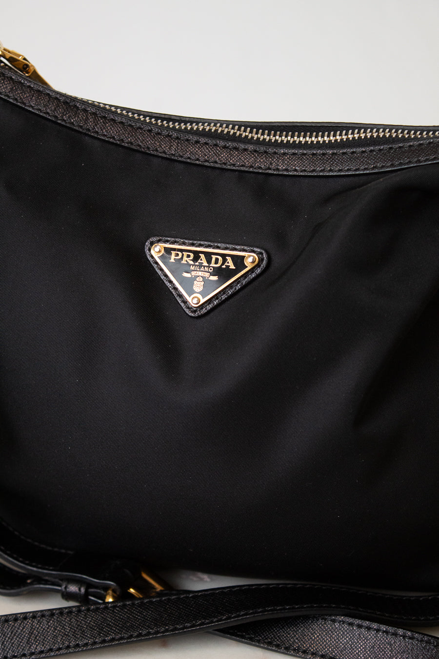 Close-up of a black Prada nylon bag with the iconic triangular Prada logo plaque in gold-tone metal on the front, showing a portion of the black zipper and strap against a light gray background.