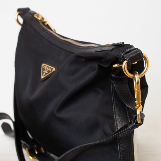 A side close- up of a black Prada nylon bag. With triangular gold hardware brand logo stating PRADA, with a zipper closure and adjustable black strap.