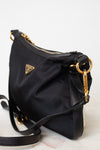 A side close- up of a black Prada nylon bag. With triangular gold hardware brand logo stating PRADA, with a zipper closure and adjustable black strap.