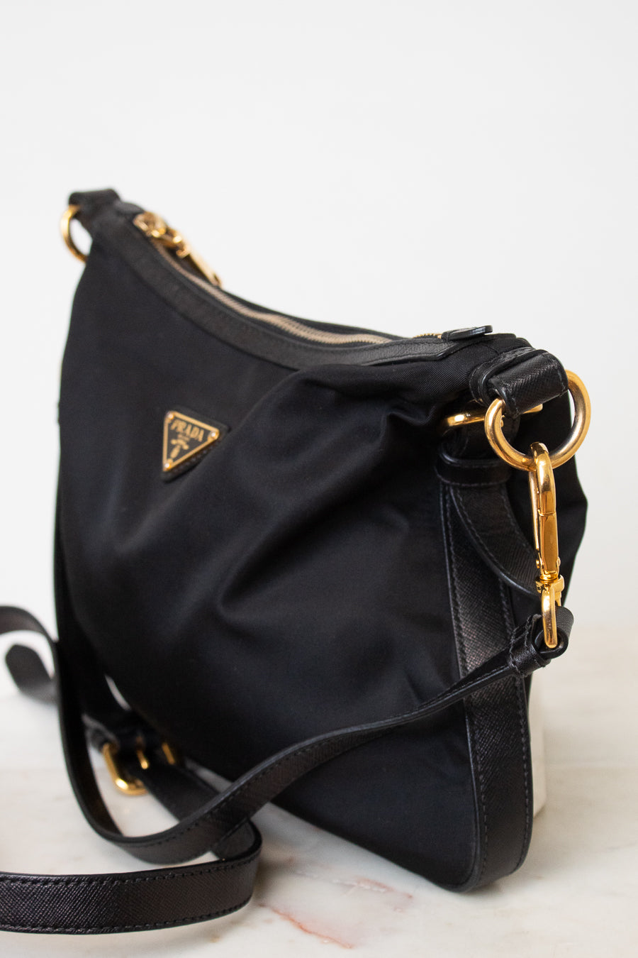 A side close- up of a black Prada nylon bag. With triangular gold hardware brand logo stating PRADA, with a zipper closure and adjustable black strap.