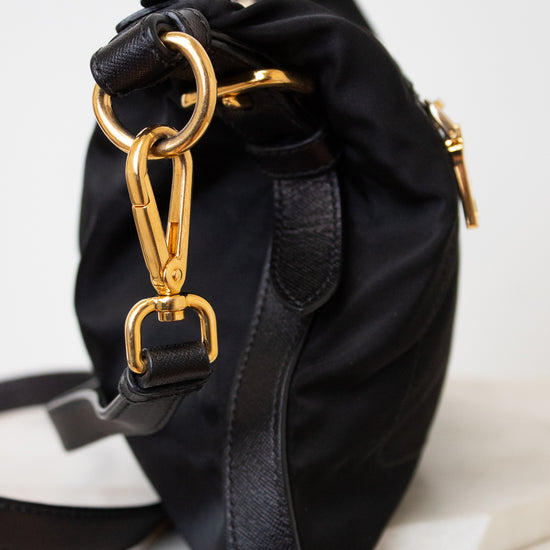 A black nylon shoulder bag with gold-tone hardware, featuring a drawstring top, leather trim, and a detachable shoulder strap, resting on a white marble surface.