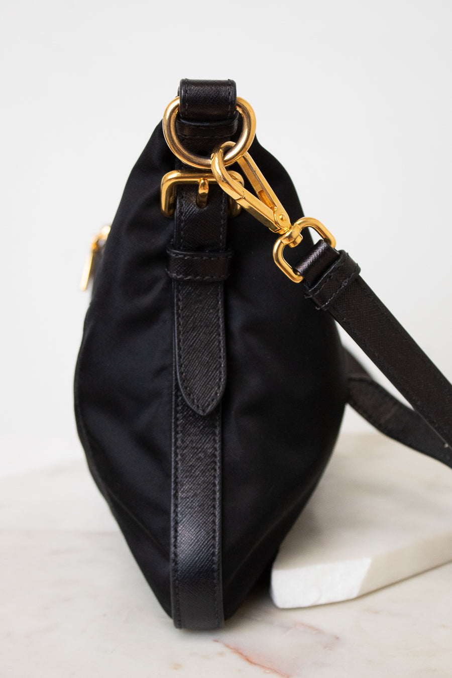 Side view of a black PRADA nylon shoulder bag featuring gold-tone ring hardware, a gold chain detail, stitched side panels , and a adjustable black shoulder strap.