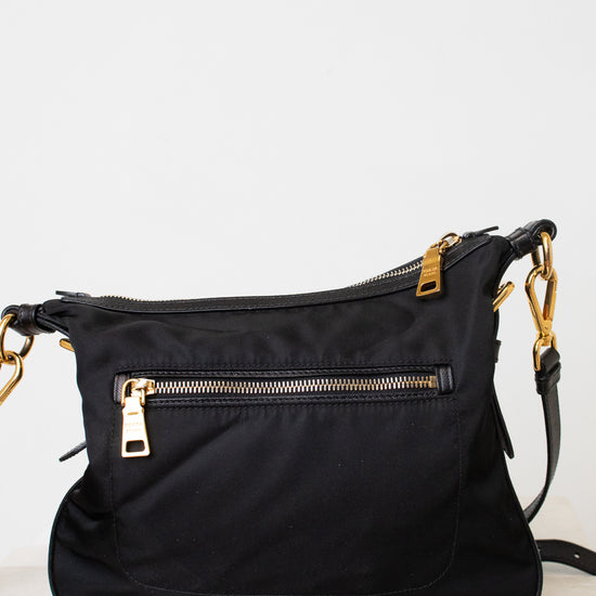 A black nylon shoulder bag with gold-tone hardware, featuring a front zippered pocket and a top zip closure, resting on a light marble surface against a white wall.