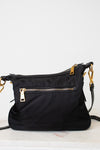 A black nylon shoulder bag with gold-tone hardware, featuring a front zippered pocket and a top zip closure, resting on a light marble surface against a white wall.