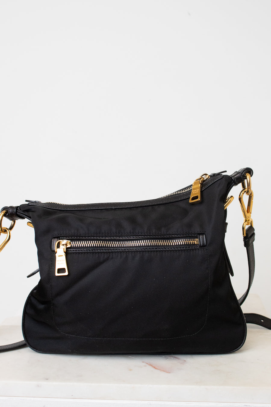 A black nylon shoulder bag with gold-tone hardware, featuring a front zippered pocket and a top zip closure, resting on a light marble surface against a white wall.
