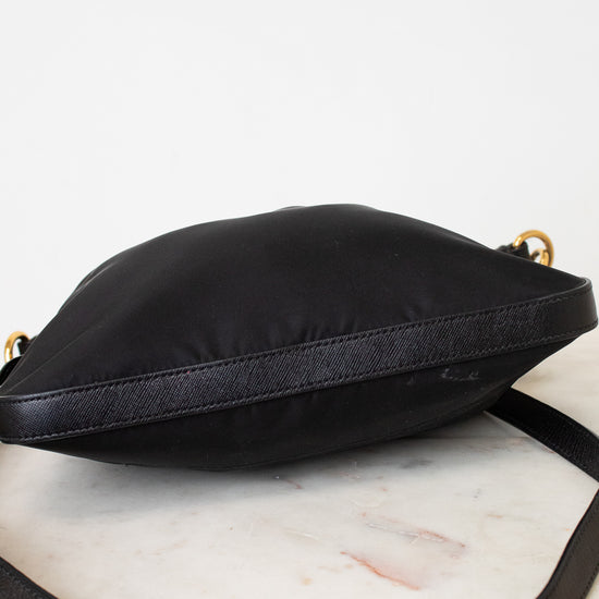 A black nylon crossbody bag with a black leather strap and gold-tone hardware, resting on a white marble surface against a white background.