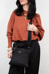 Person wearing a rust-colored sweater and black pants, holding a black Prada bag against a white background.
