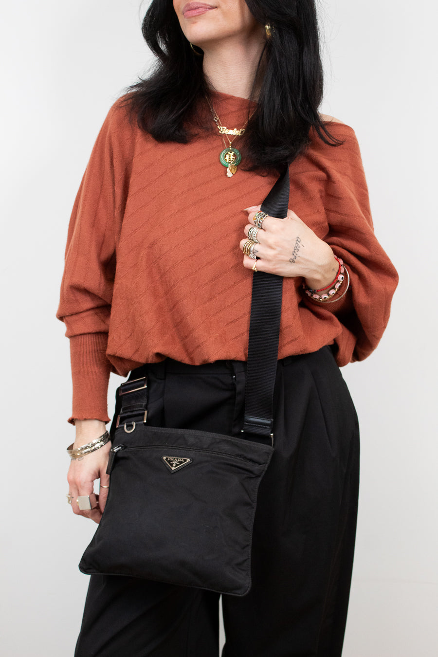 Person wearing a rust-colored sweater and black pants, holding a black Prada bag against a white background.