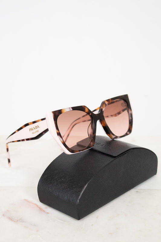 Prada sunglasses with a tortoiseshell pattern, light pink frames, and pink-tinted lenses, displayed on a black leather case against a white background.