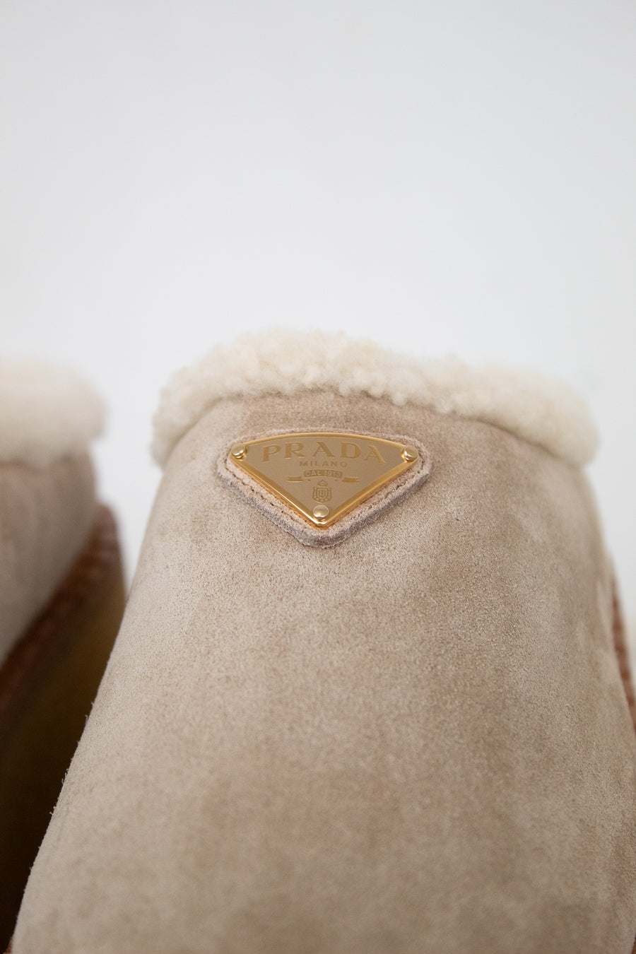 Close-up of beige suede and shearling slippers with a gold brand label on a white background