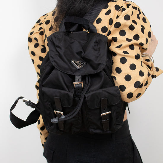 Woman wearing polka dot top and black pants holding 'PRADA' backpack on one shoulder and standing against a plain background