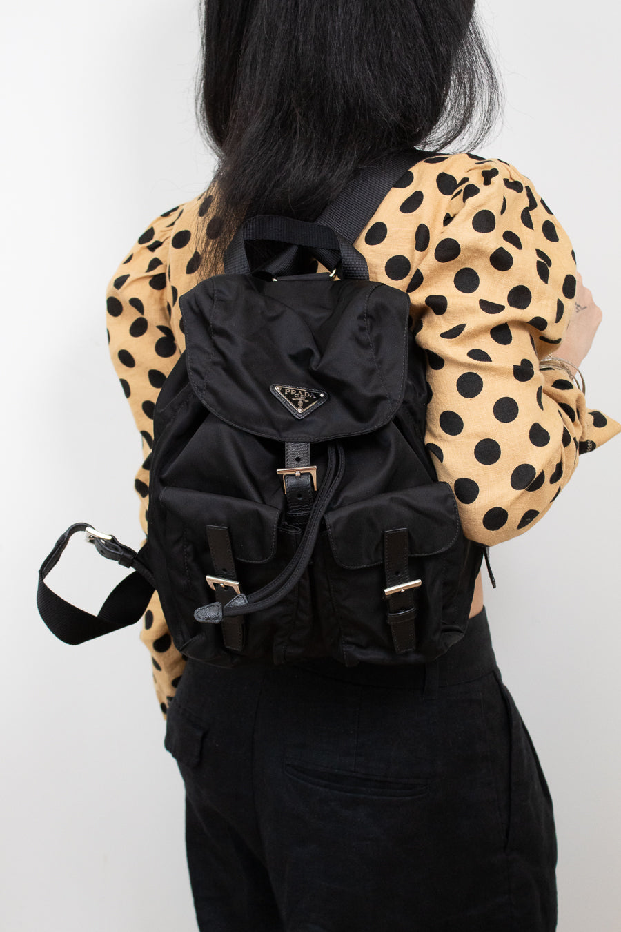 Woman wearing polka dot top and black pants holding 'PRADA' backpack on one shoulder and standing against a plain background