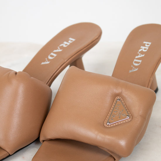 A pair of Prada tan leather slide mules with a triangular logo plaque on the strap, resting on a light marble surface against a white background.