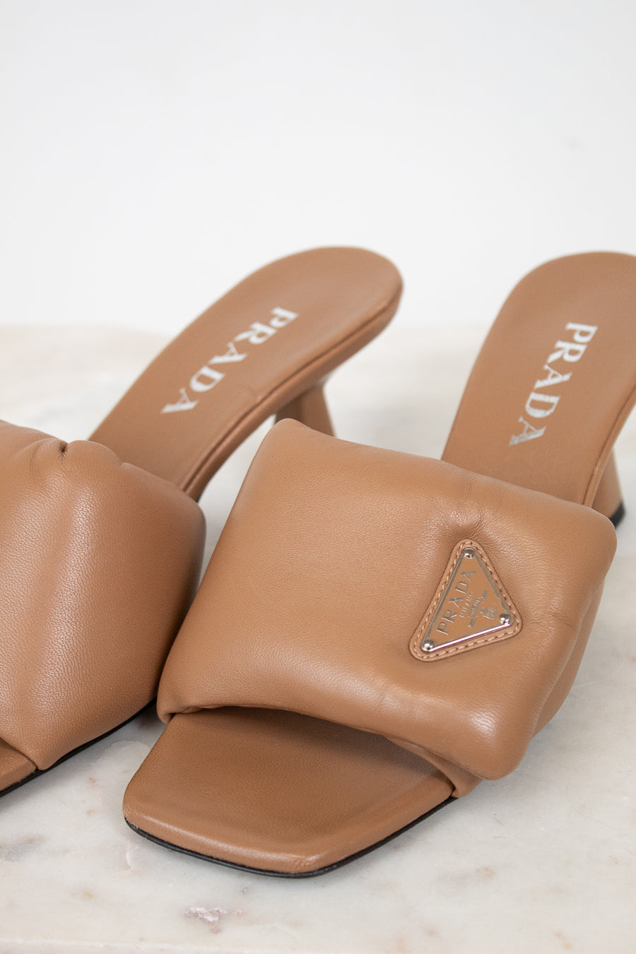 A pair of Prada tan leather slide mules with a triangular logo plaque on the strap, resting on a light marble surface against a white background.
