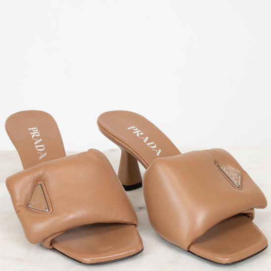 A pair of Prada tan leather slide mule heels with a geometric strap design and a small rhinestone-embellished triangle logo on the toe, displayed on a light marble surface against a white background.