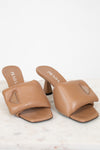 A pair of Prada tan leather slide mule heels with a geometric strap design and a small rhinestone-embellished triangle logo on the toe, displayed on a light marble surface against a white background.