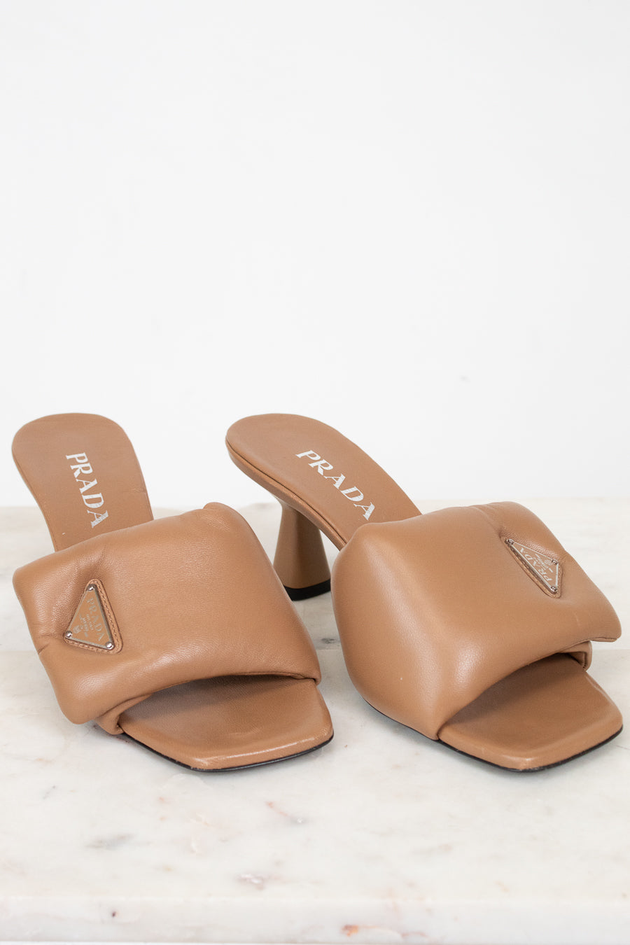 A pair of Prada tan leather slide mule heels with a geometric strap design and a small rhinestone-embellished triangle logo on the toe, displayed on a light marble surface against a white background.