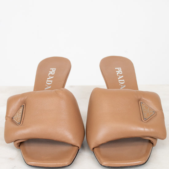 A pair of Prada tan leather slide sandals with geometric straps and a small triangular logo plaque on the side, displayed on a light marble surface against a white background.