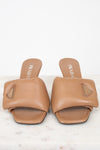 A pair of Prada tan leather slide sandals with geometric straps and a small triangular logo plaque on the side, displayed on a light marble surface against a white background.