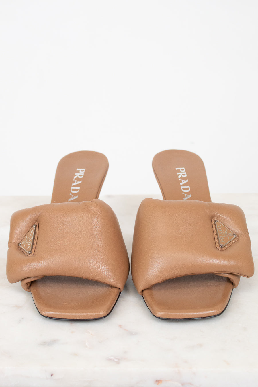 A pair of Prada tan leather slide sandals with geometric straps and a small triangular logo plaque on the side, displayed on a light marble surface against a white background.