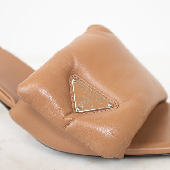 A close-up of a tan leather slide sandal featuring a prominent triangular logo plaque on the strap, set against a clean white background.