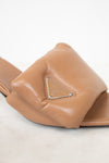 A close-up of a tan leather slide sandal featuring a prominent triangular logo plaque on the strap, set against a clean white background.