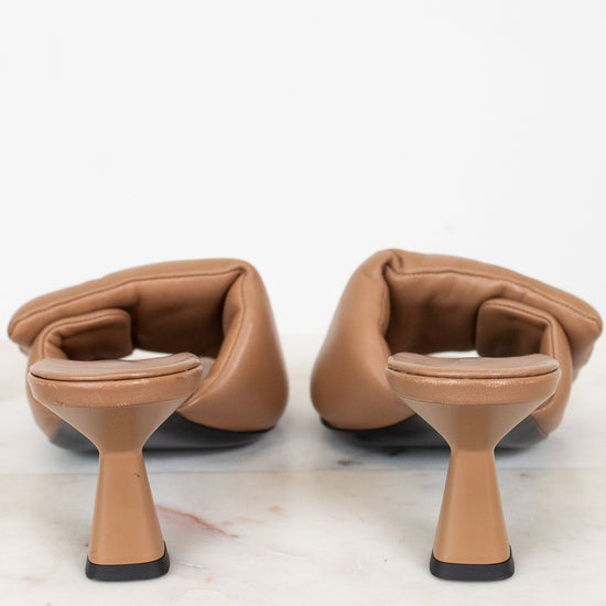 A pair of tan leather mule heels with a unique flared block heel and open toe, shown from the back on a white marble surface against a white background.