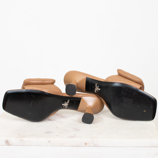A pair of women's heeled sandals shown from the bottom, featuring black square soles with a small silver logo and tan leather straps and block heels, resting on a white marble slab against a white background.