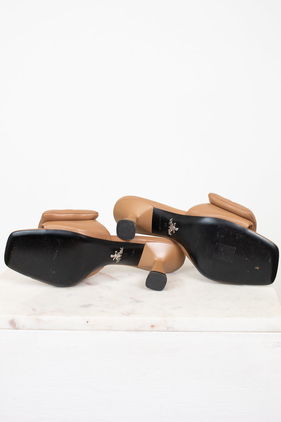 A pair of women's heeled sandals shown from the bottom, featuring black square soles with a small silver logo and tan leather straps and block heels, resting on a white marble slab against a white background.