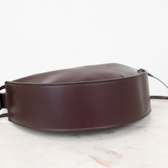 A dark burgundy leather shoulder bag with a wide, structured strap, resting on a white marble surface against a white background.