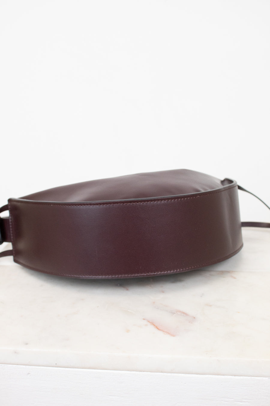 A dark burgundy leather shoulder bag with a wide, structured strap, resting on a white marble surface against a white background.