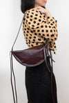 A woman wearing a tan and black polka-dot blouse and black skirt carries a dark brown crescent-shaped leather shoulder bag with a thin brown strap against a plain white background.