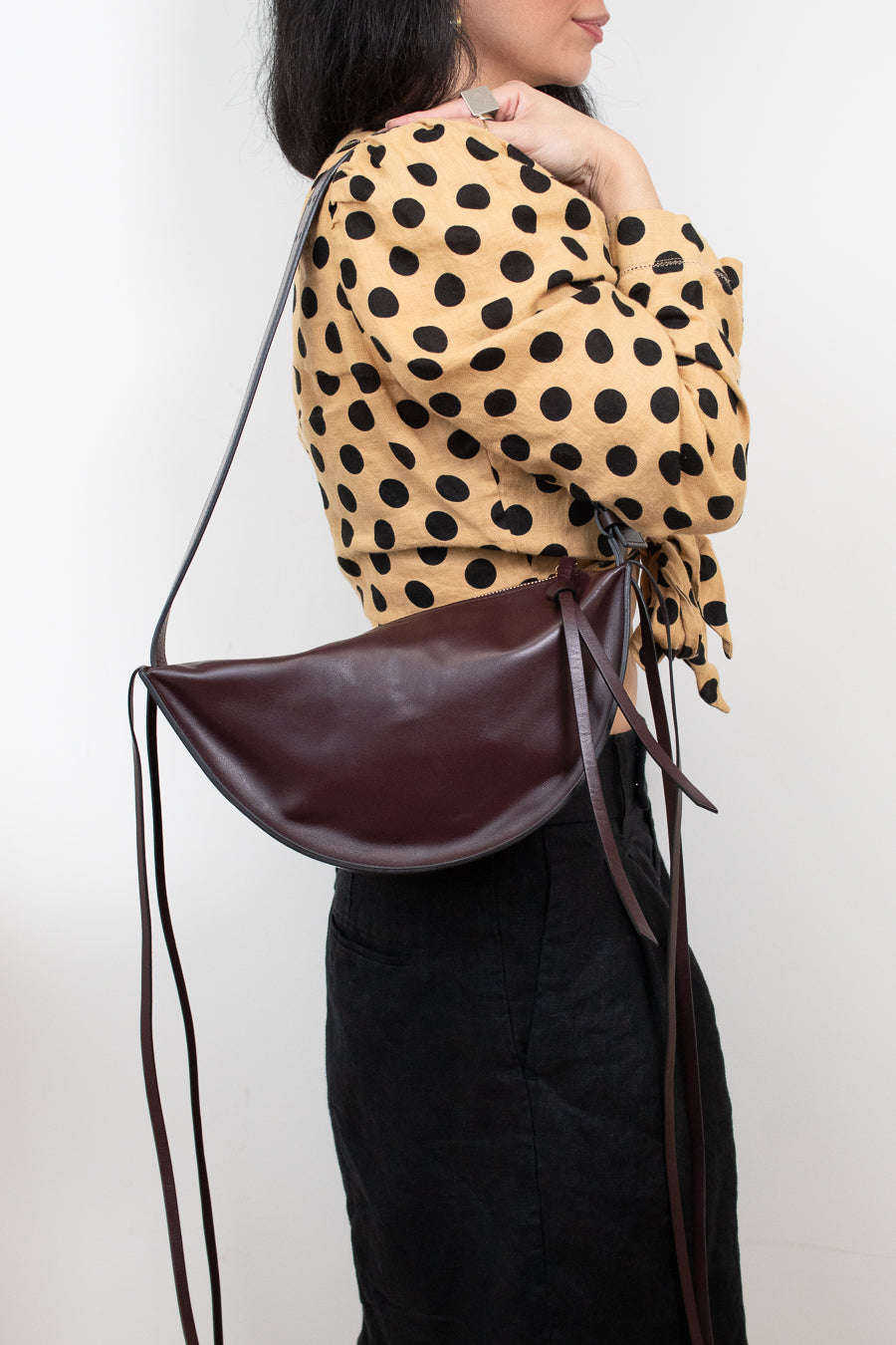 A woman wearing a tan and black polka-dot blouse and black skirt carries a dark brown crescent-shaped leather shoulder bag with a thin brown strap against a plain white background.
