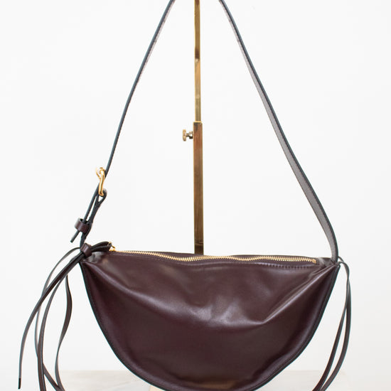 A burgundy leather crescent-shaped shoulder bag with a long thin strap and gold-tone hardware, displayed on a brass stand against a white background.