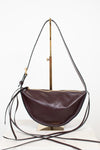 A burgundy leather crescent-shaped shoulder bag with a long thin strap and gold-tone hardware, displayed on a brass stand against a white background.
