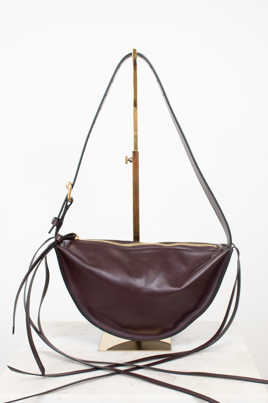 A burgundy leather crescent-shaped shoulder bag with a long thin strap and gold-tone hardware, displayed on a brass stand against a white background.