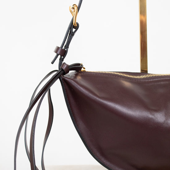 A close-up of a dark brown leather crescent-shaped shoulder bag with a gold zipper, long thin leather straps, and gold-tone hardware, displayed on a stand against a white background.