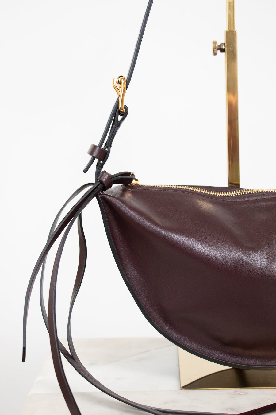 A close-up of a dark brown leather crescent-shaped shoulder bag with a gold zipper, long thin leather straps, and gold-tone hardware, displayed on a stand against a white background.