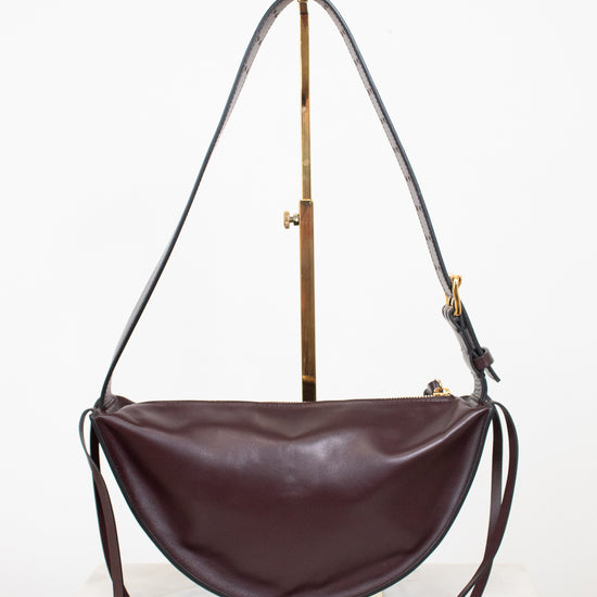 A dark burgundy leather crescent-shaped shoulder bag with a long thin strap, displayed on a gold-colored stand against a white background.
