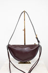 A dark burgundy leather crescent-shaped shoulder bag with a long thin strap, displayed on a gold-colored stand against a white background.